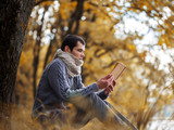 A young man in a warm knitted sweater and scarf Snood reading an old book in the autumn Park . Lifestyle