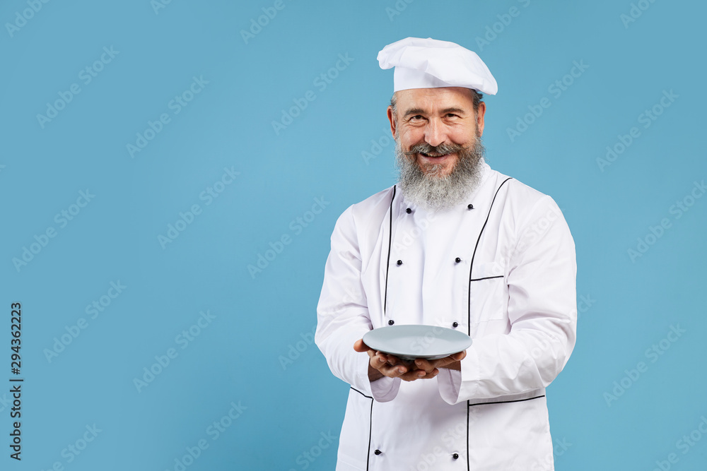 Portrait of bearded senior chef holding empty plate and smiling at camera while standing against blue background, mock up template with copy space Portrait of bearded senior chef holding empty plate and smiling at camera while standing against blue background, mock up template with copy space
