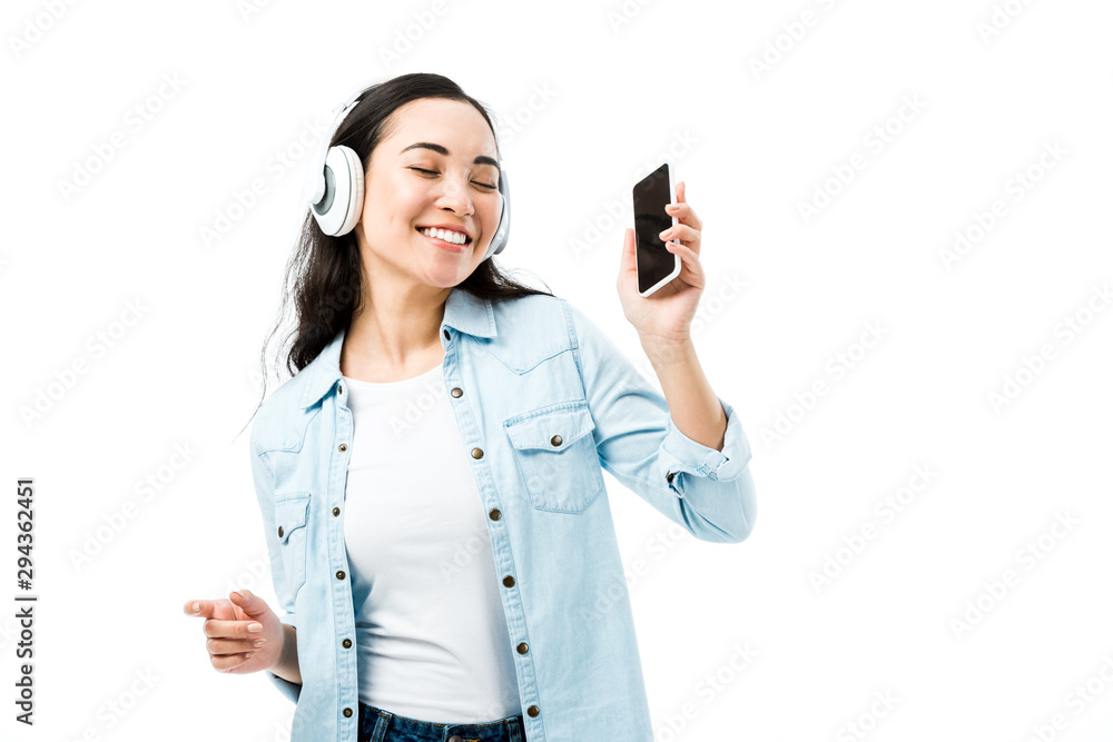 attractive and smiling asian woman in denim shirt listening music and holding smartphone isolated on white attractive and smiling asian woman in denim shirt listening music and holding smartphone isolated on white