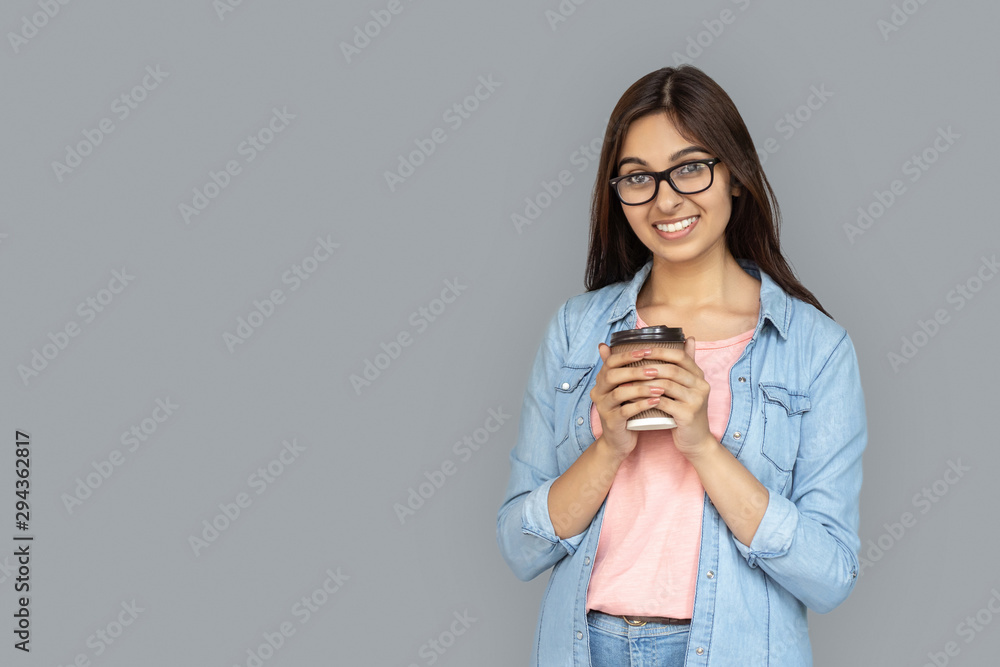 Happy and cheerful young adult woman standing isolated on grey background with copy space. Smiling indian girl looking at camera and holding paper cup with beverage in hands Happy and cheerful young adult woman standing isolated on grey background with copy space. Smiling indian girl looking at camera and holding paper cup with beverage in hands