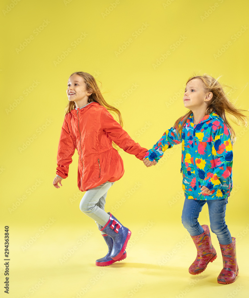 A full length portrait of a bright fashionable girls in a raincoat holding hands, running and having fun on yellow studio background. Autumn and spring fashion for kids. Cute stylish blonde girls. A full length portrait of a bright fashionable girls in a raincoat holding hands, running and having fun on yellow studio background. Autumn and spring fashion for kids. Cute stylish blonde girls.