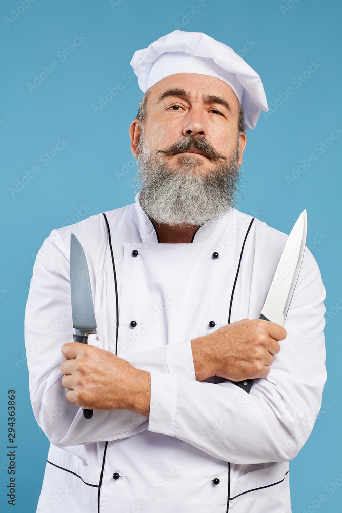 Waist up portrait of charismatic bearded chef looking at camera holding two knives while standing against blue background Waist up portrait of charismatic bearded chef looking at camera holding two knives while standing against blue background