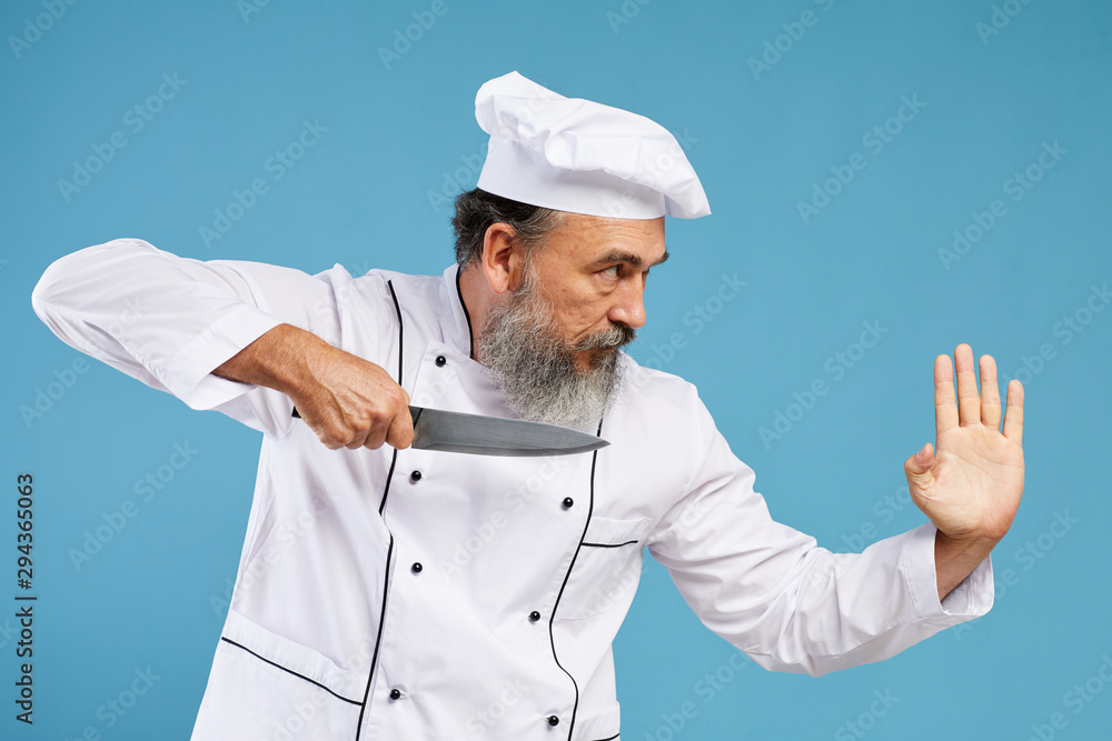 Side view portrait of charismatic bearded chef holding sharp knife standing en garde while posing against blue background in studio, copy space Side view portrait of charismatic bearded chef holding sharp knife standing en garde while posing against blue background in studio, copy space