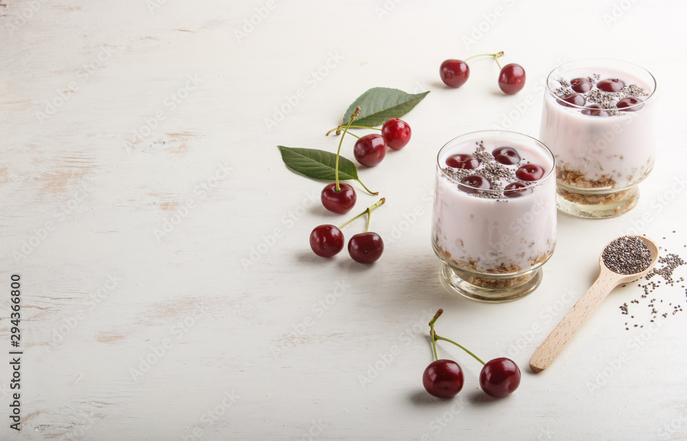 Yoghurt with cherries, chia seeds and granola in glass with wooden spoon on white wooden background. side  view.