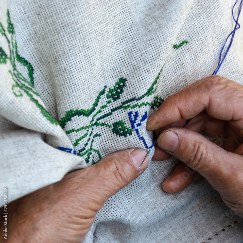Hands of an elderly woman embroidering a cross-stitch floral pattern on linen fabric. Embroidery, handwork, needlecraft concept. Closeup