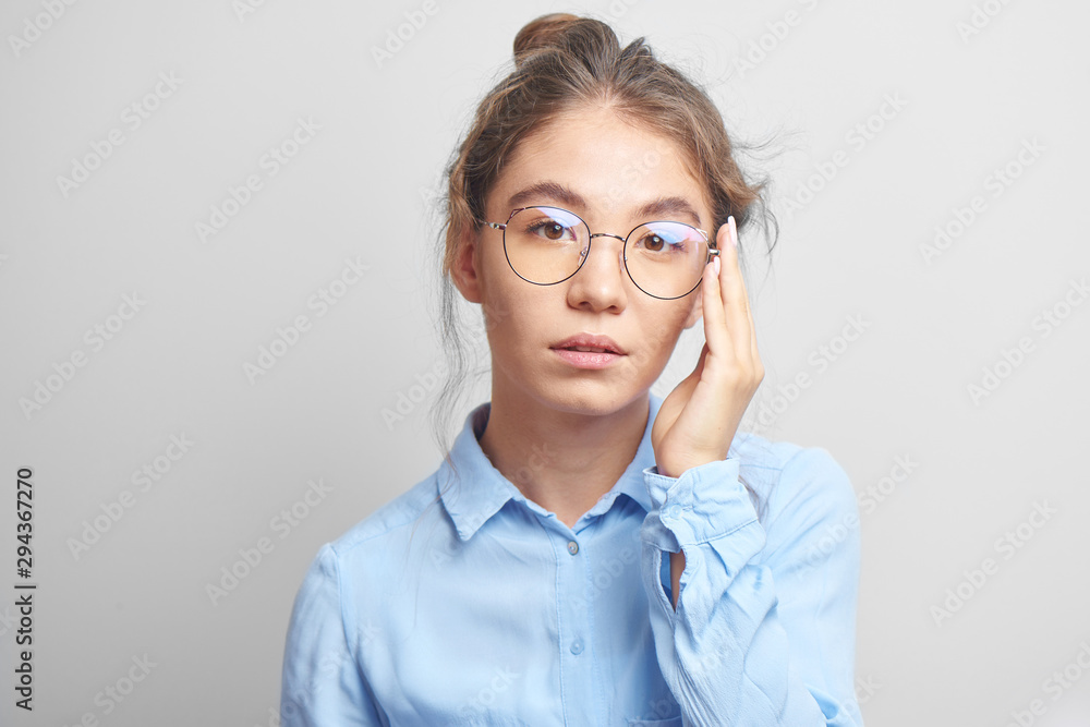 Portrait of pensive Asian Kazakh young woman with bun of hair on head in stylish glasses wearing a shirt in white studio, thinking girl Portrait of pensive Asian Kazakh young woman with bun of hair on head in stylish glasses wearing a shirt in white studio, thinking girl