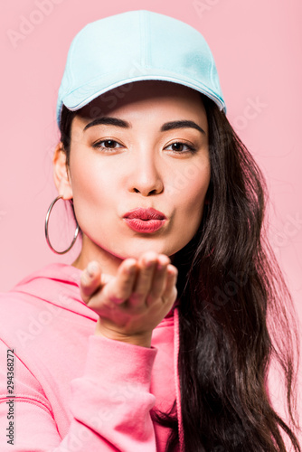 attractive asian woman in pink pullover and cap sending air kiss isolated on pink