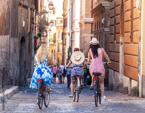 Three happy young women friends tourists with bikes in typical alley in Rome, Italy at sunrise.
