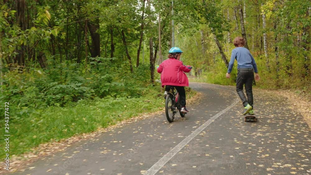 Back view of children riding bicycle and skateboard in park. Rear view of two cute boys riding bike and longboard in beautiful park Back view of children riding bicycle and skateboard in park. Rear view of two cute boys riding bike and longboard in beautiful park