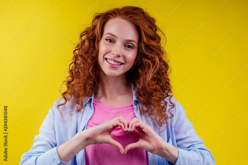 happy young redhaired ginger woman with curly hairstyle pretty face wearing  cotton pink shirt ,making heart gesture with hands symbol in studio yellow  background Stock Photo | Adobe Stock