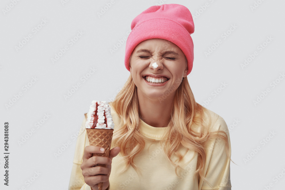 Attractive young woman eating ice cream and smiling while standing against grey background Attractive young woman eating ice cream and smiling while standing against grey background