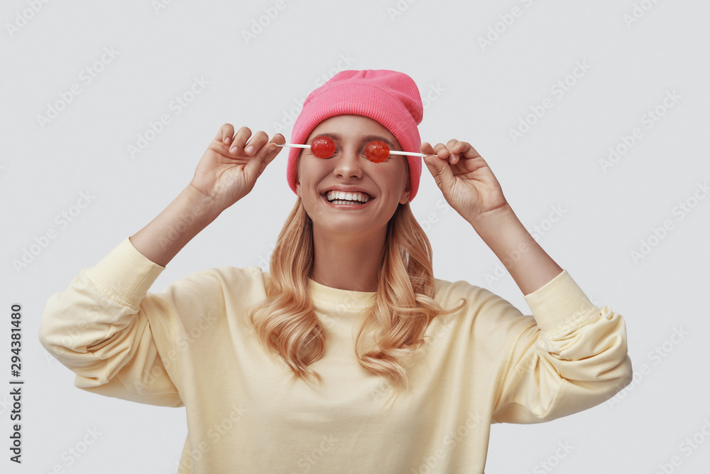 Attractive young woman smiling and covering eyes with lollipops while standing against grey background Attractive young woman smiling and covering eyes with lollipops while standing against grey background