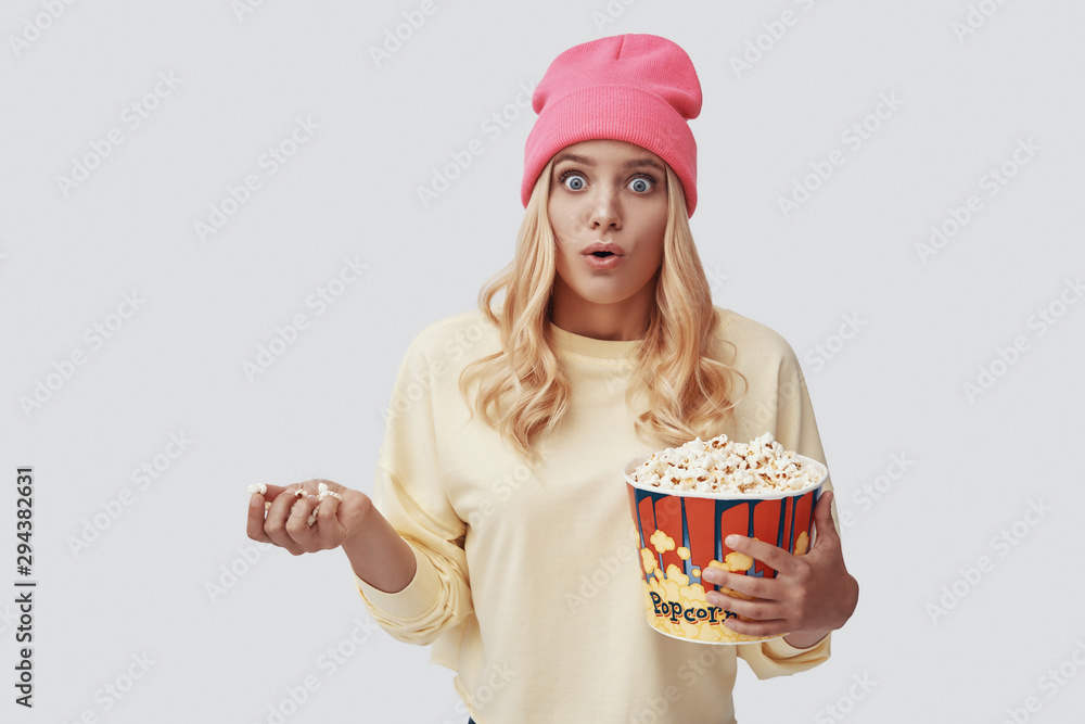 Attractive young woman eating popcorn and making a face while standing against grey background Attractive young woman eating popcorn and making a face while standing against grey background
