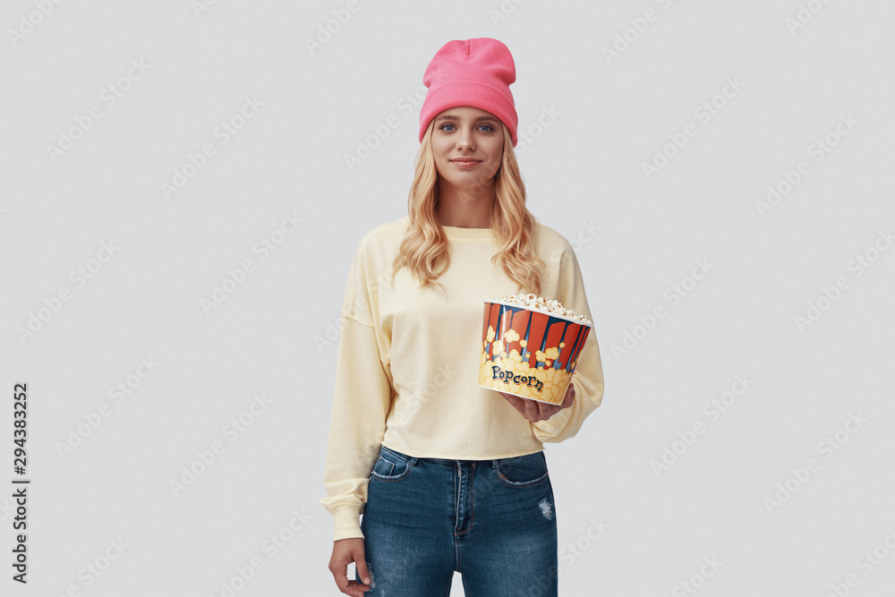 Attractive young woman holding popcorn and smiling while standing against grey background Attractive young woman holding popcorn and smiling while standing against grey background