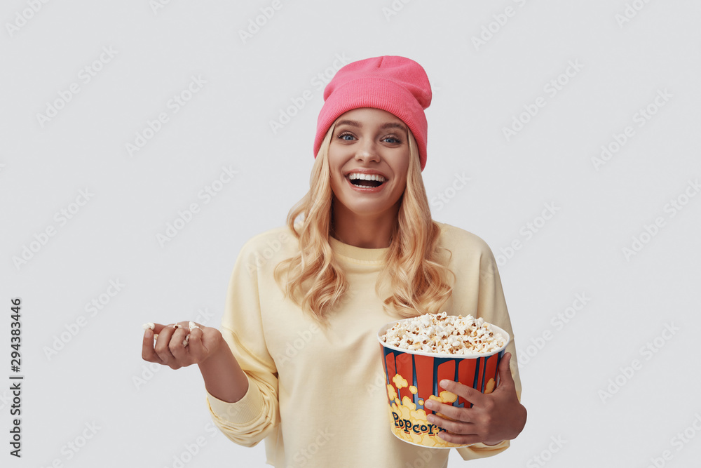 Attractive young woman eating popcorn and smiling while standing against grey background Attractive young woman eating popcorn and smiling while standing against grey background