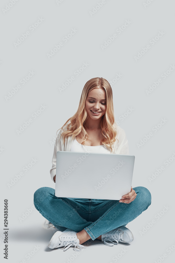 Naklejka premium Attractive young woman using laptop and smiling while sitting against grey background
