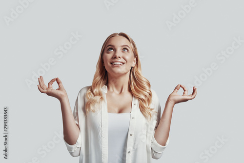 Beautiful young woman meditating and smiling while standing against grey background
