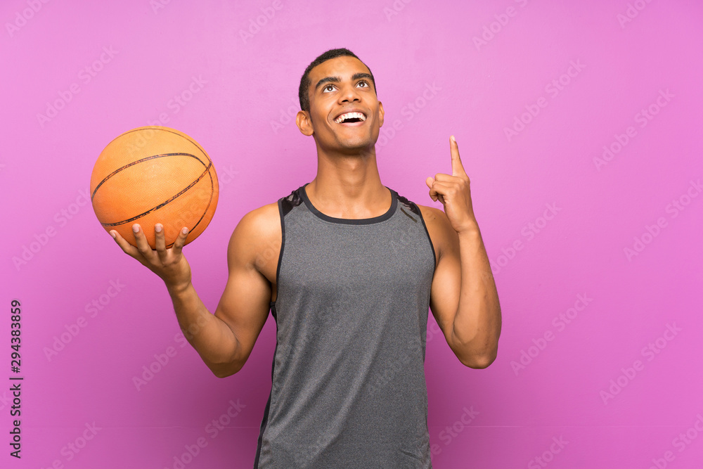 Young sport man with ball of basketball over isolated purple wall pointing up a great idea