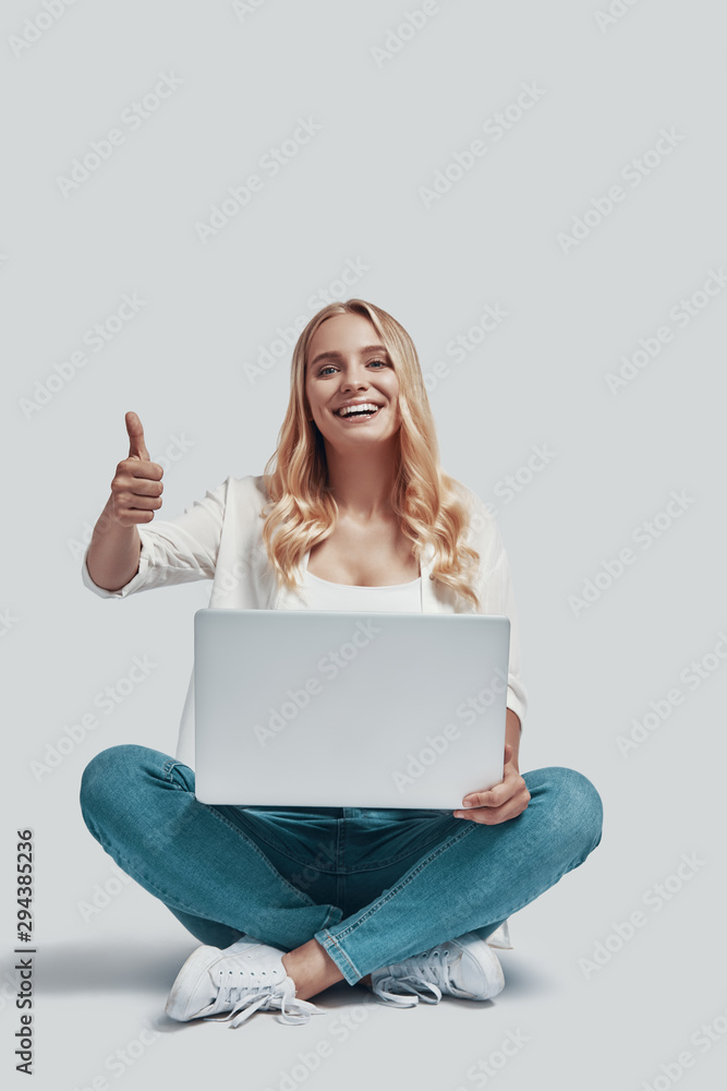 Attractive young woman using laptop and smiling while sitting against grey background Attractive young woman using laptop and smiling while sitting against grey background