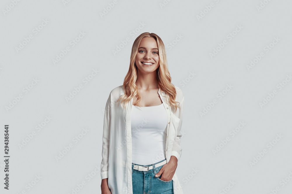 Attractive young woman looking at camera and smiling while standing against grey background Attractive young woman looking at camera and smiling while standing against grey background