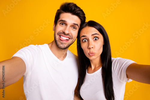 Photo of two people in foolish mood taking selfies sticking tongue making faces wear casual clothes isolated yellow color background