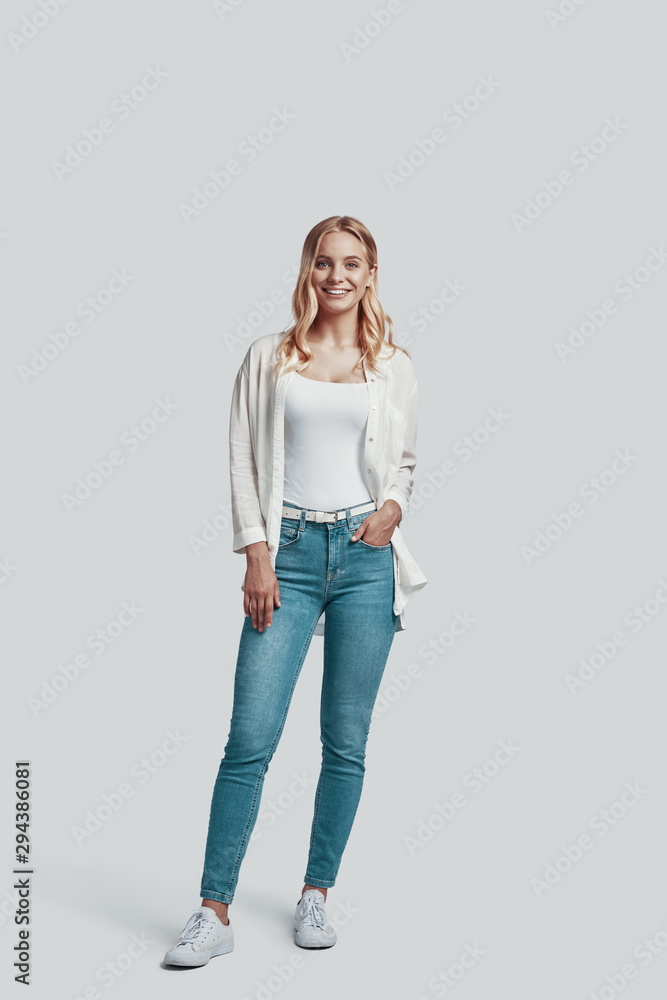 Full length of attractive young woman looking at camera and smiling while standing against grey background Full length of attractive young woman looking at camera and smiling while standing against grey background