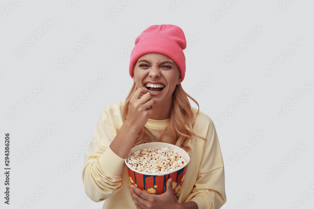 Attractive young woman eating popcorn and smiling while standing against grey background Attractive young woman eating popcorn and smiling while standing against grey background