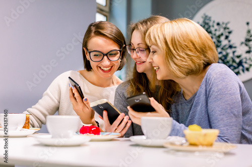 Image of three excited happy pretty girls friends sitting in cafe  drinking coffee  eating cakes and using phones.