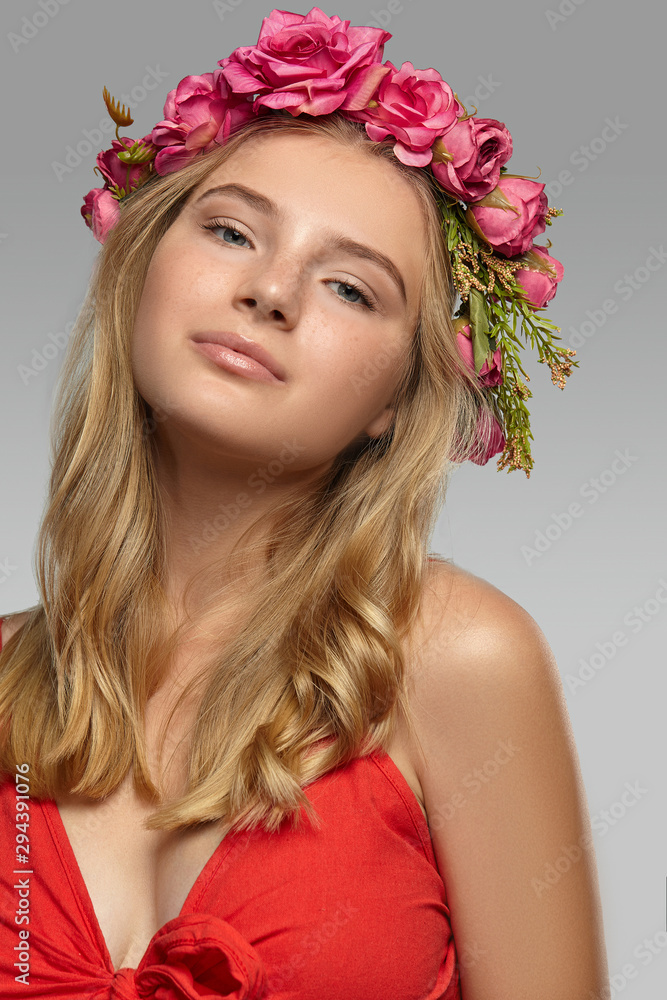 Close-up shot of a romantic blonde European lady with wavy hair in a fire-red low necked top and a delicate circlet made of crimson artificial roses.  