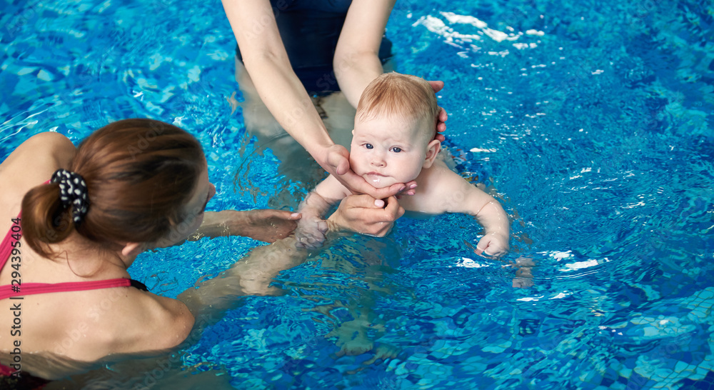 Adorable baby boy enjoying swimming in paddling turquoise pool with his mother and instructor. Child swimming trainer in action. Sport and healthy development from first year of life. Cropped view