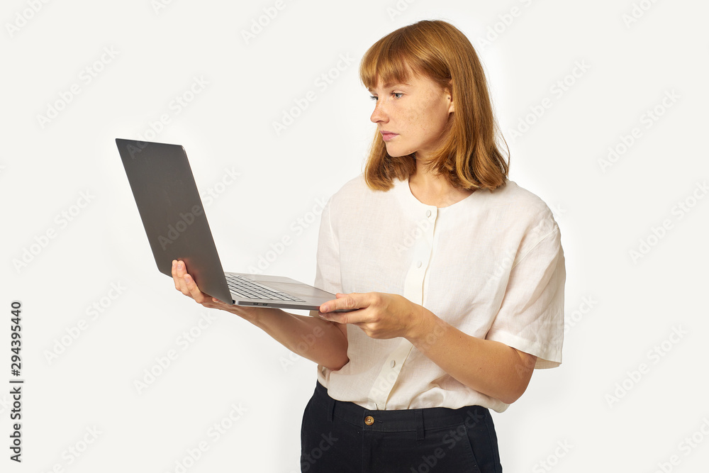 Headshot of young woman with red hair and freckles holding laptop and looking at the screen working. Isolated on white background with copyspace