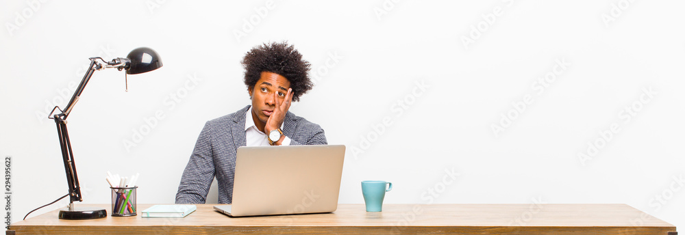 © kues1 - young black businessman feeling bored, frustrated and sleepy after a tiresome, dull and tedious task, holding face with hand on a desk