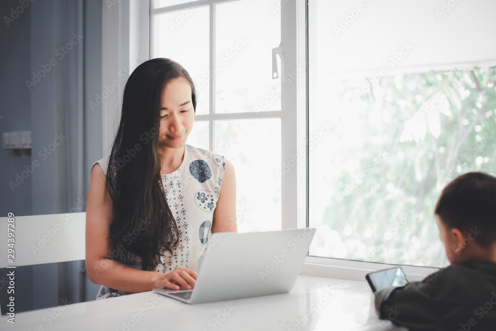 Beautiful Asian girls using laptop computer and her son looking at smartphone. Raise children and work concept. Beautiful Asian girls using laptop computer and her son looking at smartphone. Raise children and work concept.
