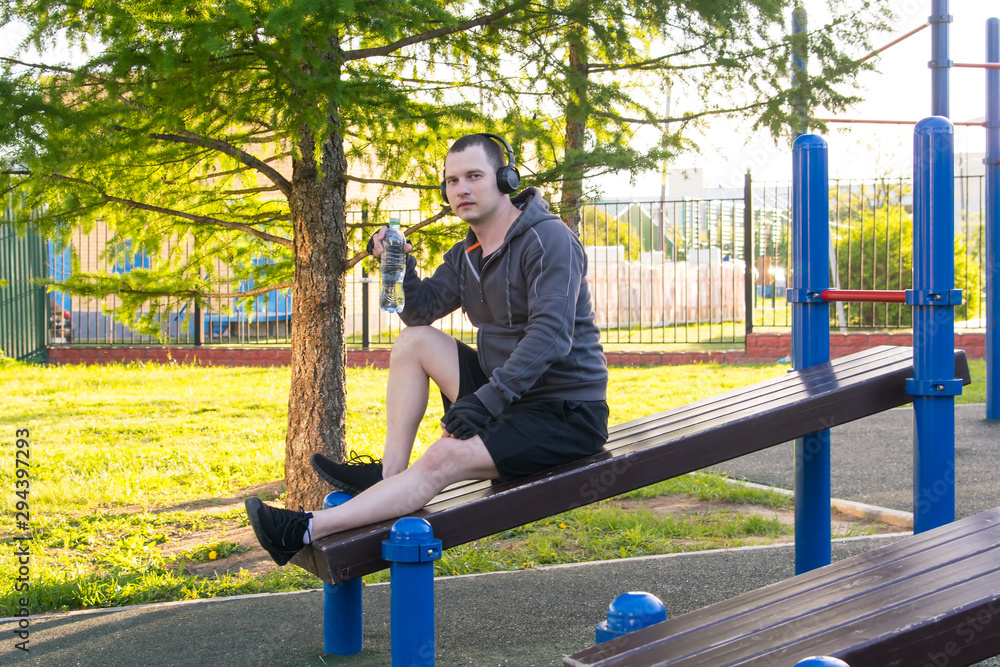 between intervals of sports approaches, the man holds a bottle of water and listens to music, sitting on the simulator, outdoors