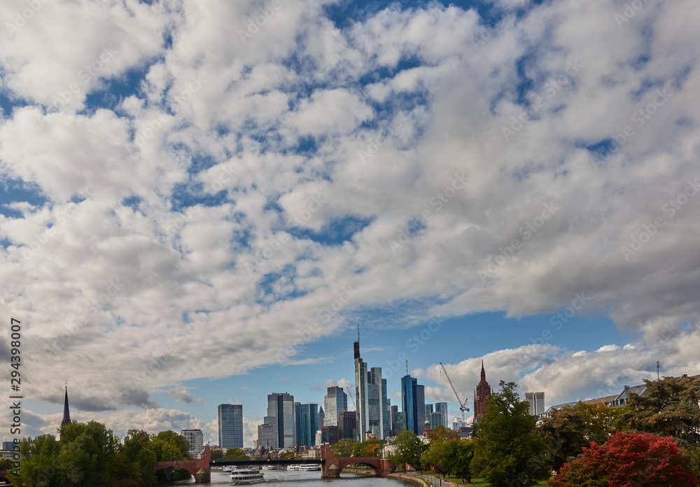 Skyline with the skyscrapers of Frankfurt and the river Main in the foreground with big cloudy blue and white sky Skyline with the skyscrapers of Frankfurt and the river Main in the foreground with big cloudy blue and white sky