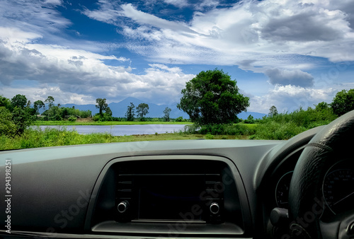 Beatuful landscape view from inside car. Steering wheel and dashboard of car interior. Road trip travel with scenic view of mountain, lake, and forest. Blue sky and white fluffy clouds. Vacation time.