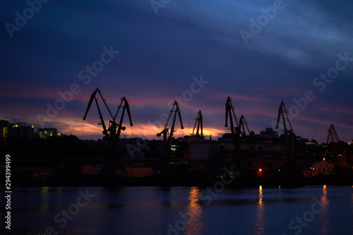 Colorful sunset over sea port and industrial cranes. Bright sunset in seaport. Large silhouettes of cargo cranes. Beautiful landscape with fiery sunset sky and sea.