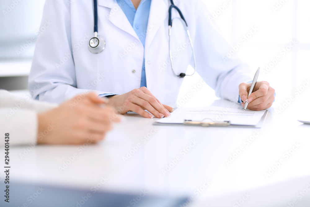 Woman doctor and patient sitting and talking at medical examination at hospital office, close-up. Physician filling up medication history records. Medicine and healthcare concept Woman doctor and patient sitting and talking at medical examination at hospital office, close-up. Physician filling up medication history records. Medicine and healthcare concept