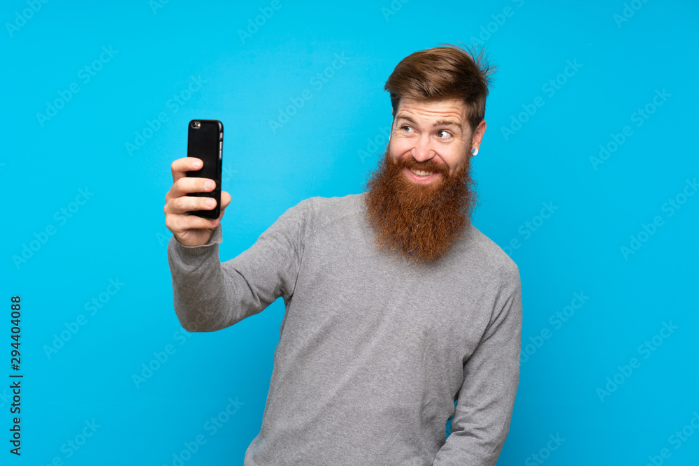 Redhead man with long beard over isolated blue background making a selfie Redhead man with long beard over isolated blue background making a selfie