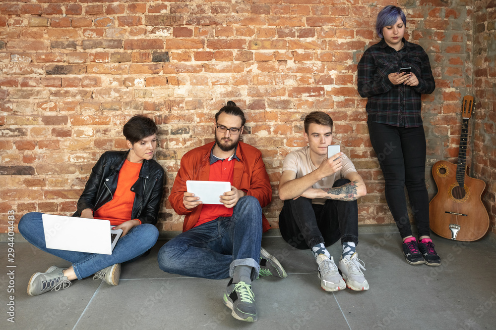 Group of happy caucasian young people behind the brick wall. Sharing a news, photos or videos from smartphones, laptops or tablets, playing games and having fun. Social media, modern technologies.