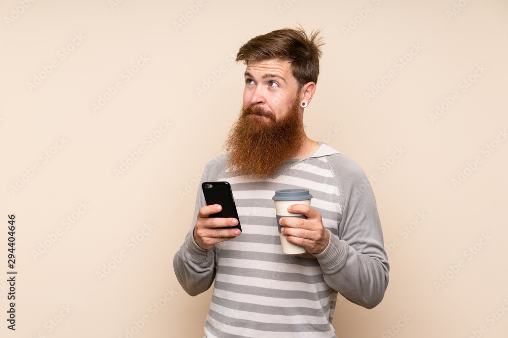 Redhead man with long beard over isolated background holding coffee to take away and a mobile while thinking something Redhead man with long beard over isolated background holding coffee to take away and a mobile while thinking something