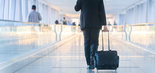 Rear view of unrecognizable formaly dressed businessman walking and wheeling a trolley suitcase at the lobby, talking on a mobile phone. Business travel concept.