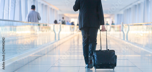 Rear view of unrecognizable formaly dressed businessman walking and wheeling a trolley suitcase at the lobby, talking on a mobile phone. Business travel concept.