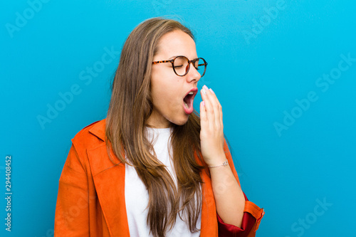 young woman yawning lazily early in the morning, waking and looking sleepy, tired and bored against blue background
