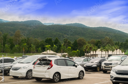 Car parking in large asphalt parking lot with trees, white cloud and blue sky mountain background