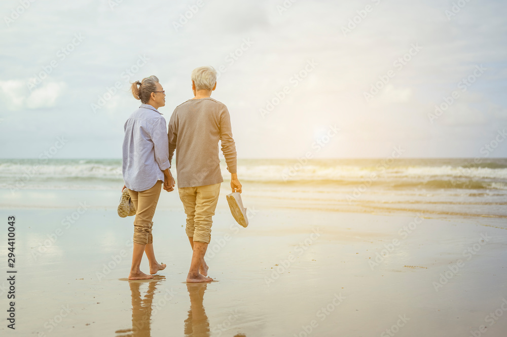 Senior couple walking on the beach holding hands at sunrise, plan life insurance at retirement concept. - obrazy, fototapety, plakaty 