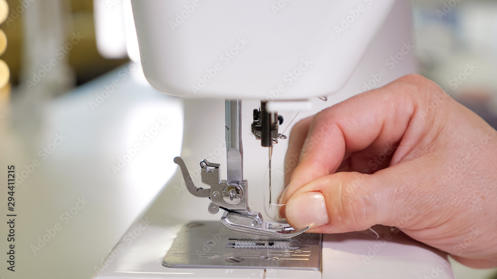 Tailoring business concept. Woman puts thread in needle in sewing machine preparing to work, hands closeup. Dressmaker sews on sewing mashine.