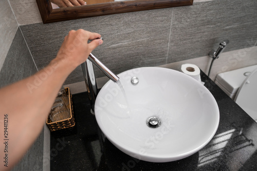 Male hand use a faucet in a bathroom interior with white round sink and chrome faucet. Water flowing from the chrome faucet.