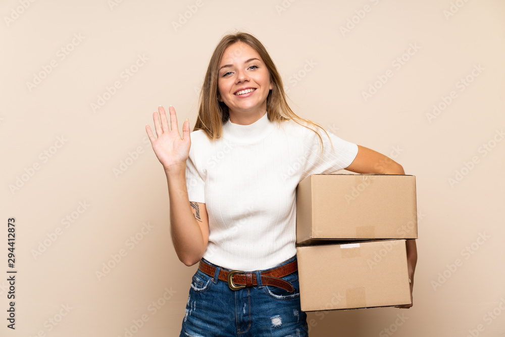 Young blonde woman over isolated background holding a box to move it to another site and saluting Young blonde woman over isolated background holding a box to move it to another site and saluting