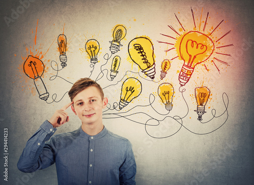 Confident teenage boy pointing forefinger to head showing where genius ideas come from, and shining light bulbs sketches like different thoughts. Genius creativity concept, planning way to success.